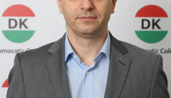 A portrait of Nyakó István, a Hungarian politician, standing confidently in front of a backdrop featuring the DK (Democratic Coalition) party logo and Hungarian political symbols, with a clear, professional appearance and a serious yet approachable expression, captured in high detail