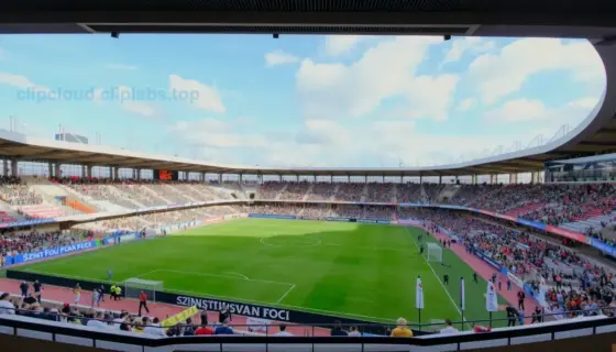 A wide-angle view of a modern football stadium terrace with a large banner displaying 'Szinva Foci' and another banner showing 'Szent István Foci', both set against a clear sky, featuring fans in team colors, goalposts, and a lush green pitch, captured in a realistic style with high detail and vibrant colors