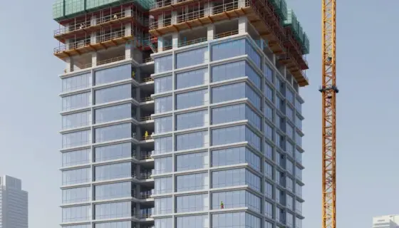 A modern architectural rendering of the AVALON BUSINESS CENTER being constructed, showing the building's exterior with glass facade, steel framework, and construction cranes, with workers in safety gear visible on scaffolding, set against a clear daytime sky with city skyline in the background, emphasizing the development progress of the business center