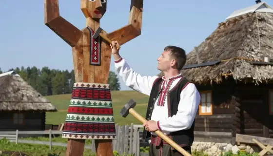 A young Hungarian boy in traditional folk attire (embroidered shirt, waistcoat, trousers) stands proudly next to a large, ornate wooden Rád (a traditional Hungarian folk art figure) in a rural Hungarian village setting. The boy holds a small wooden tool, possibly a hammer or chisel, as if he has just finished building or repairing the Rád. In the background, there is a rustic wooden house with a thatched roof, green fields, and a clear blue sky. The scene is bright and sunny, conveying a sense o