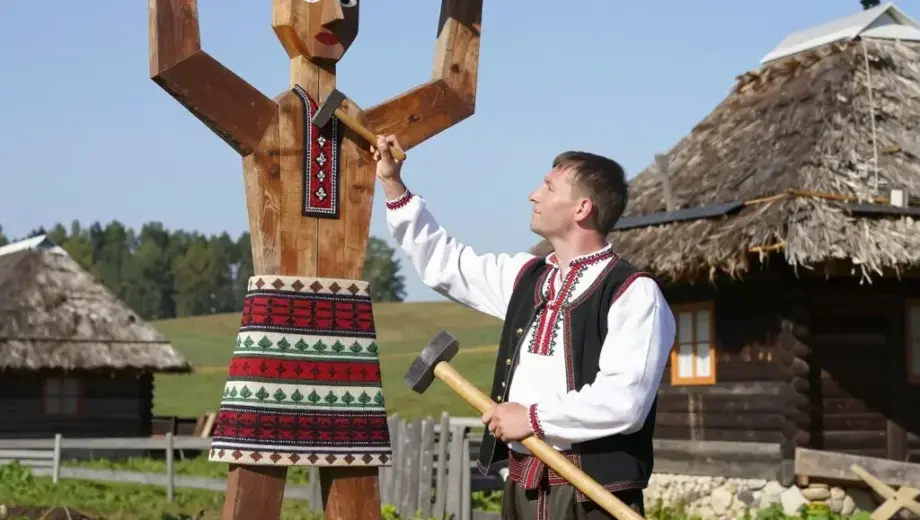 A young Hungarian boy in traditional folk attire (embroidered shirt, waistcoat, trousers) stands proudly next to a large, ornate wooden Rád (a traditional Hungarian folk art figure) in a rural Hungarian village setting. The boy holds a small wooden tool, possibly a hammer or chisel, as if he has just finished building or repairing the Rád. In the background, there is a rustic wooden house with a thatched roof, green fields, and a clear blue sky. The scene is bright and sunny, conveying a sense o
