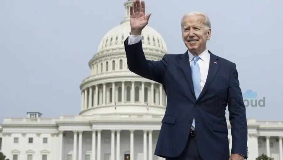 Joe Biden standing in front of the United States Capitol building, smiling and waving, with American flags in the background, clear sky, formal attire