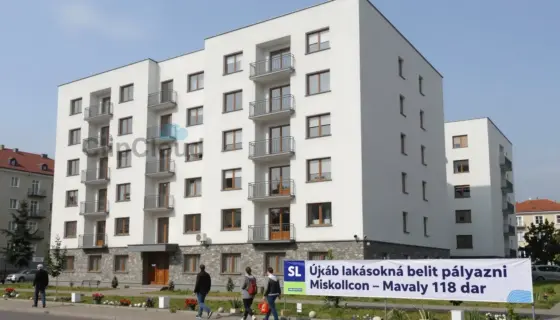 A modern residential building in Miskolc with several apartments visible, people walking by, a banner or sign indicating 'Újabb lakásokra lehet pályázni Miskolcon – tavaly 118 dar' in Hungarian, urban setting with trees and clear sky, realistic and detailed depiction of housing opportunity