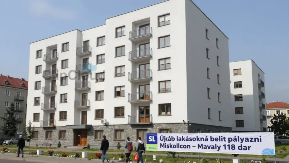 A modern residential building in Miskolc with several apartments visible, people walking by, a banner or sign indicating 'Újabb lakásokra lehet pályázni Miskolcon – tavaly 118 dar' in Hungarian, urban setting with trees and clear sky, realistic and detailed depiction of housing opportunity