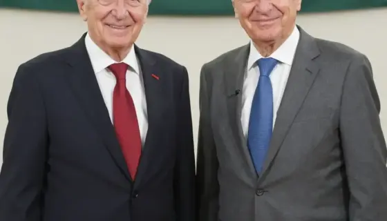 A formal portrait of two elderly Hungarian men, László Kövér and Péter Polt, celebrating their 50th anniversary. László Kövér is on the left, wearing a dark suit with a red tie, smiling warmly. Péter Polt is on the right, wearing a grey suit with a blue tie, also smiling. They are standing in front of a backdrop with the Hungarian flag and a banner that reads '50 éves'. The scene is well-lit, with a professional studio setting, capturing a moment of celebration and respect.