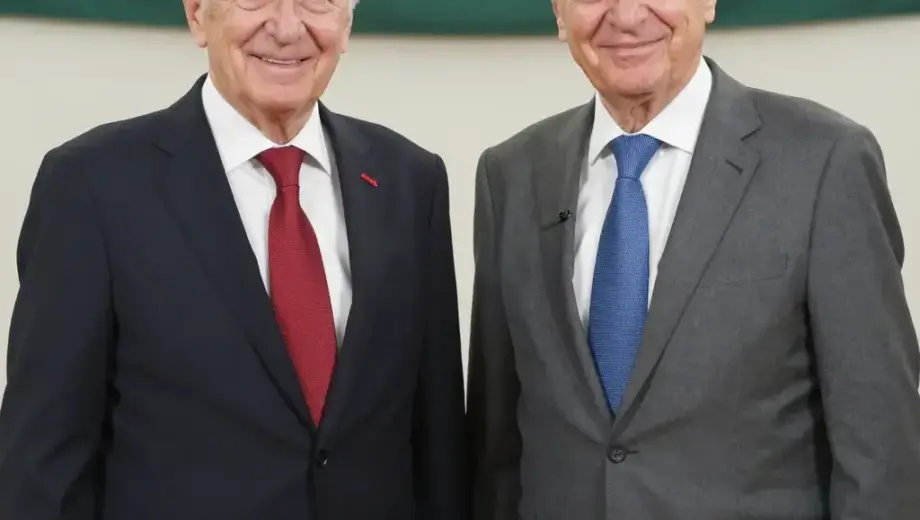 A formal portrait of two elderly Hungarian men, László Kövér and Péter Polt, celebrating their 50th anniversary. László Kövér is on the left, wearing a dark suit with a red tie, smiling warmly. Péter Polt is on the right, wearing a grey suit with a blue tie, also smiling. They are standing in front of a backdrop with the Hungarian flag and a banner that reads '50 éves'. The scene is well-lit, with a professional studio setting, capturing a moment of celebration and respect.