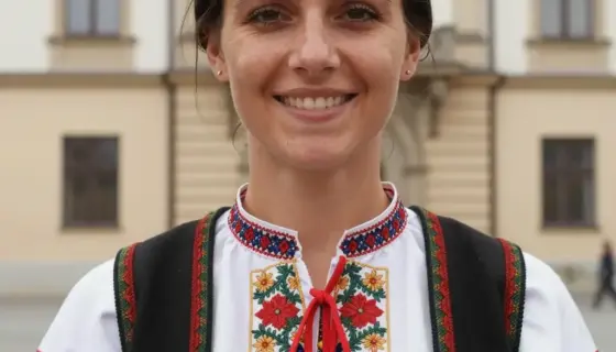 A portrait of a woman with dark hair, wearing a traditional Hungarian folk costume, standing in front of a historic building in Miskolc, Hungary, with a warm and welcoming expression, natural lighting, detailed facial features, and a clear view of the building's architecture