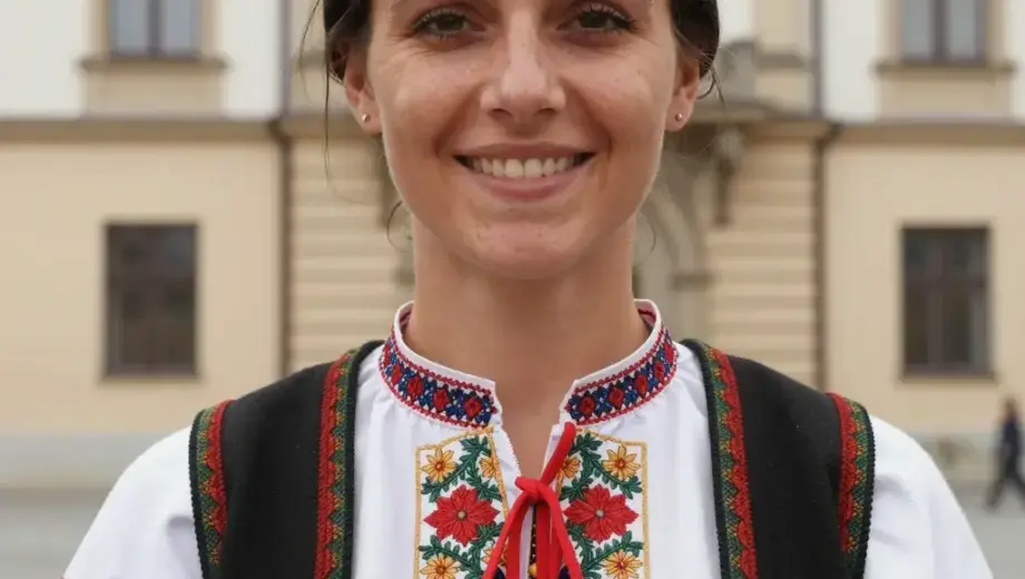 A portrait of a woman with dark hair, wearing a traditional Hungarian folk costume, standing in front of a historic building in Miskolc, Hungary, with a warm and welcoming expression, natural lighting, detailed facial features, and a clear view of the building's architecture