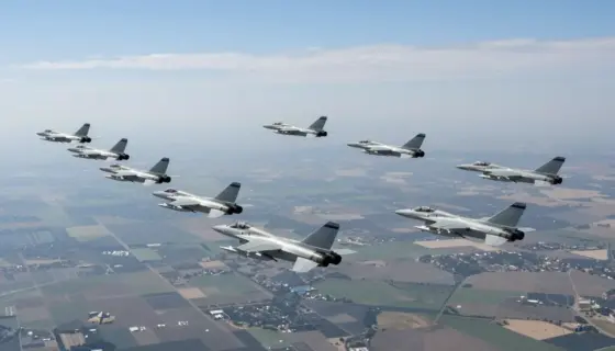 Aerial view of a large-scale military operation with approximately one hundred Israeli fighter jets flying in formation over a distant landscape, targeting long-range objectives. The scene should depict the jets in mid-flight, showcasing their precision and coordination. The background should include a clear sky with some clouds, and the distant landscape should be visible but not detailed, emphasizing the vast distance the jets are covering.