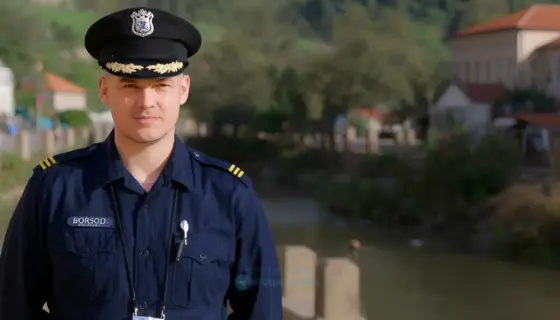 A realistic portrait of a Hungarian police officer in a traditional uniform standing in Borsod-Abaúj-Zemplén County, with a subtle transformation effect showing personal changes in their career, set in a natural outdoor environment with local landmarks like the Borsod River or historic buildings, emphasizing professional dedication and community connection