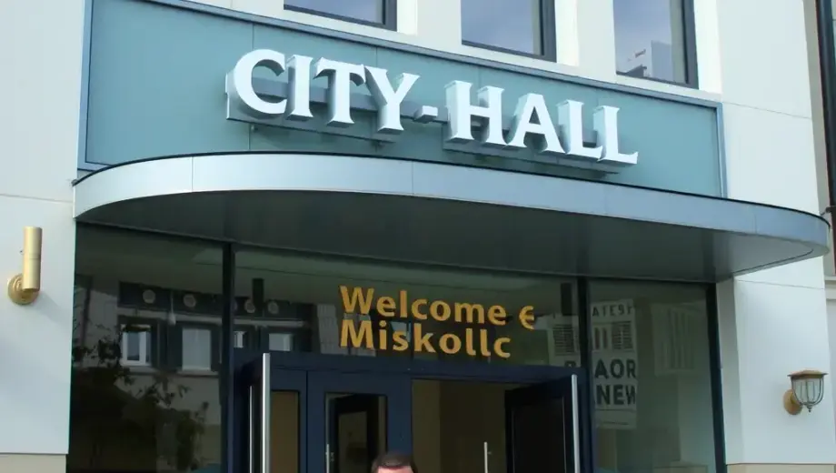 A modern city hall building in Miskolc, Hungary, with a welcoming atmosphere, showing local government officials interacting with citizens, possibly including a mayor or council members, in front of the building with clear signage indicating municipal support, daytime scene with clear visibility, architectural details of the city hall, people dressed in business casual or formal attire, symbolizing community engagement and administrative support