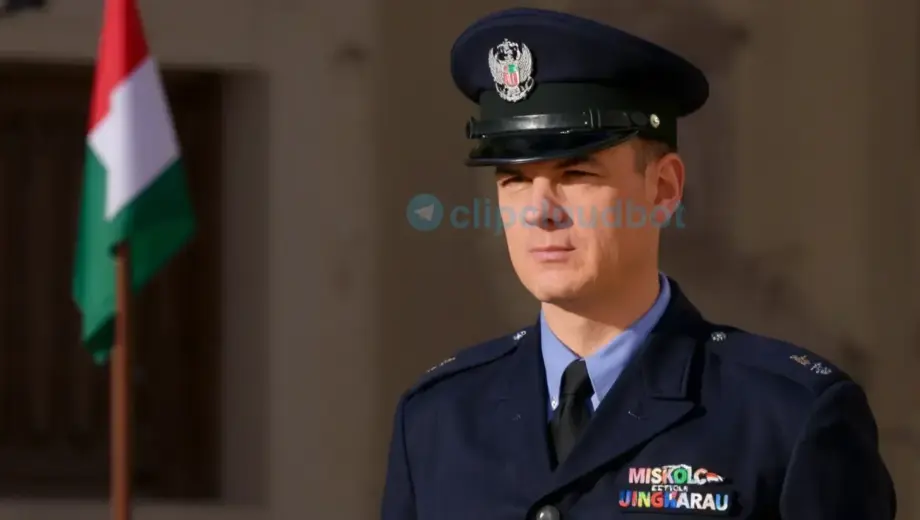 A formal portrait of a man in a dark uniform with a peaked cap, standing in front of a building with a Hungarian flag. The man has a serious expression and is looking slightly to the side. The setting appears to be an official government building, likely the Miskolc City Hall, with classical architecture visible in the background. The lighting is natural daylight, suggesting an outdoor press event or official announcement.