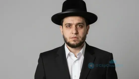 A portrait of a young man with short dark hair and a serious expression, wearing traditional Jewish religious attire including a black suit, white shirt, and black hat, standing against a plain neutral background