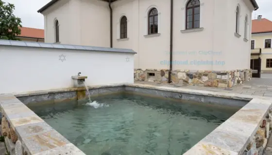 A serene Jewish ritual bath (mikveh) located next to a historic synagogue in Miskolc, Hungary. The mikveh features stone steps leading down into clear, still water, surrounded by simple, modest architecture with whitewashed walls and subtle traditional Jewish design elements. The synagogue in the background has a modest facade with arched windows and a Star of David symbol, blending into a quiet urban setting with soft daylight. No people are visible, emphasizing tranquility and reverence.