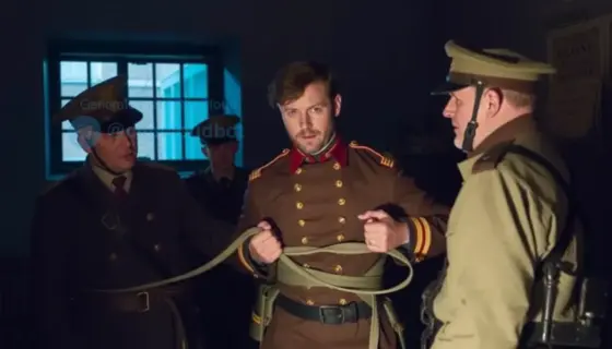 A historical scene depicting a man in 19th-century military uniform being tied up by soldiers in a dimly lit room, with a barred window and a wooden chair visible in the background, conveying a serious and tense atmosphere