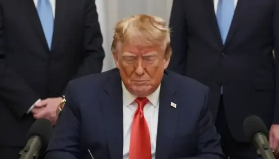 A formal indoor setting showing Donald Trump signing a document at a desk, with Hungarian officials standing nearby, microphones and cameras present, and a Hungarian flag visible in the background