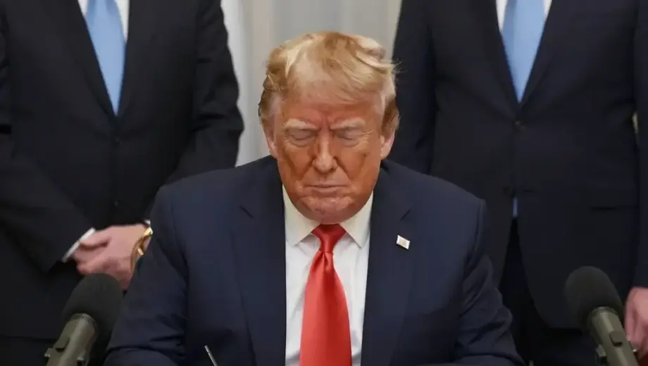 A formal indoor setting showing Donald Trump signing a document at a desk, with Hungarian officials standing nearby, microphones and cameras present, and a Hungarian flag visible in the background