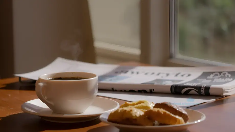 A bright, cheerful Saturday morning breakfast scene featuring a steaming cup of coffee, a plate of fresh pastries, and a newspaper on a wooden table, with sunlight streaming through a window