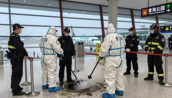 A dramatic scene at an airport terminal showing emergency responders in protective gear containing a gas leak, with a detained individual in custody nearby, and security personnel implementing safety measures such as barriers and evacuation signs, all under bright airport lighting with visible aircraft in the background through glass walls