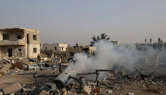 A war-torn landscape in southern Lebanon showing damaged buildings, smoke rising from rubble, and a destroyed drone wreckage on the ground, with distant silhouettes of Hezbollah fighters observing from cover, under a hazy afternoon sky, realistic war photography style