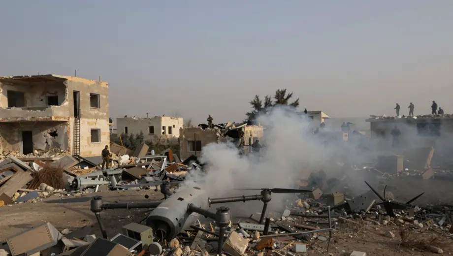 A war-torn landscape in southern Lebanon showing damaged buildings, smoke rising from rubble, and a destroyed drone wreckage on the ground, with distant silhouettes of Hezbollah fighters observing from cover, under a hazy afternoon sky, realistic war photography style